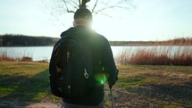 Active handsome elderly man with nordic walking sticks walks forward through along the lake at sunset. Senior man is engaged in scandinavian Walking with a backpack. Healthy lifestyle concept.