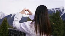 Woman making a heart shape with her hands, standing in a white bathrobe on the terrace of the hotel. Winter travelling scene, wanderlust concept.