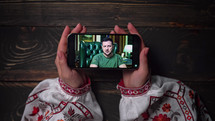August 2022 - Kyiv, Ukraine. Watching online speech of President Volodymyr Zelenskiy on smartphone on wooden table background. News from front. russian aggression.
