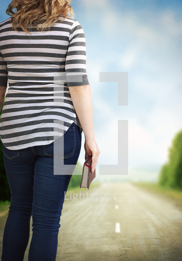 woman looking down the road ahead carrying a Bible
