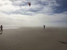Silhouette of a jogger and a man flying a kite on a beach, in the mist, near the ocean water, under the clouds.