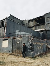 A man stands in front of an old, abandoned, industrial building.