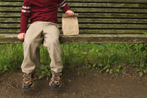 a child with a brown lunch bag sitting on a bench 