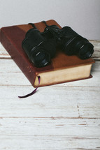 Binoculars on closed Bible laying on top of wooden table.
