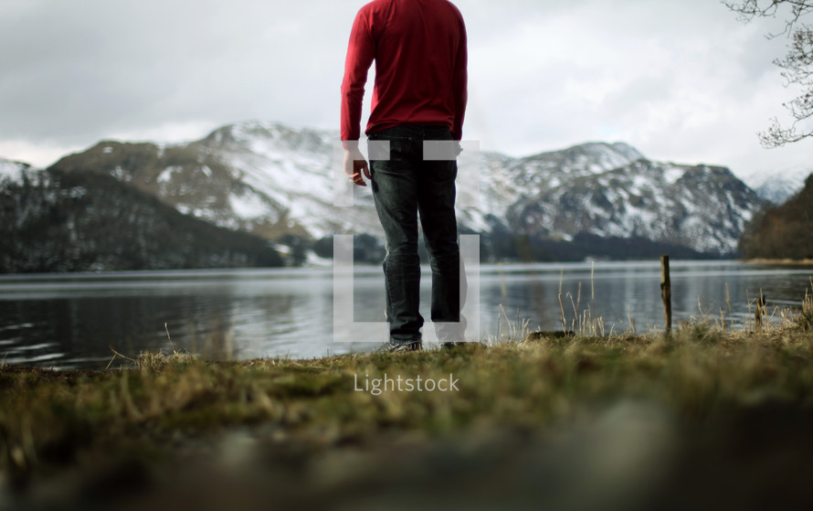 Man standing in the grass at the edge of a lake, overlooking a snow-covered mountain range.