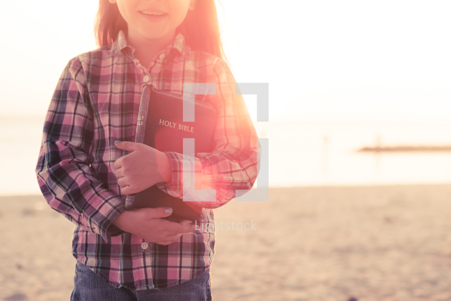 young girl happy, smiling, standing on the beach holding her bible at sunset, sunflare