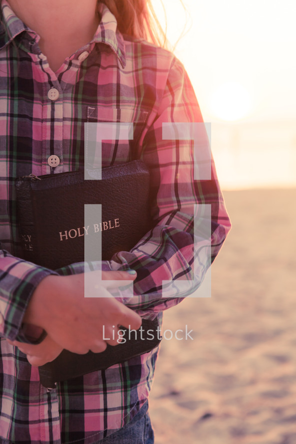 young girl standing on the beach holding her bible at sunset