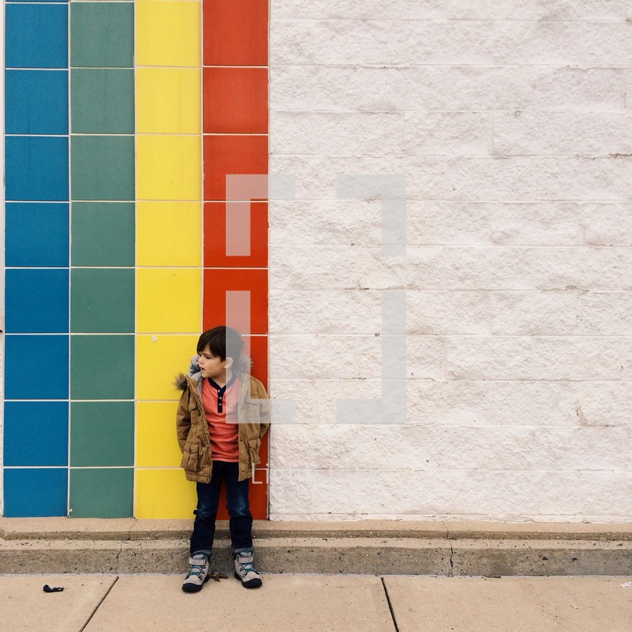 little boy standing in front of a rainbow tile wall. 