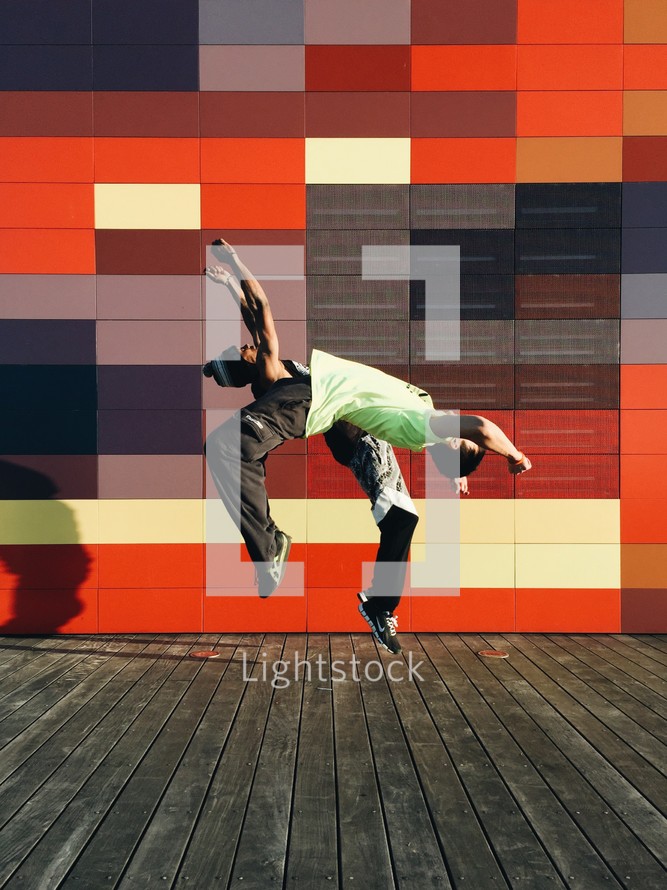 Two people doing back flips in front of a colorful wall.