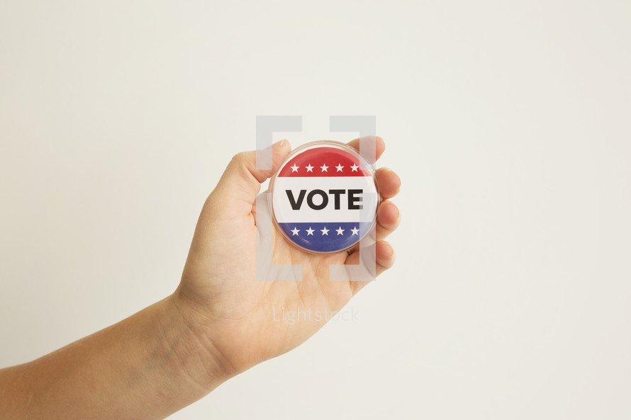 A hand holding a red white and blue button reading, "Vote."