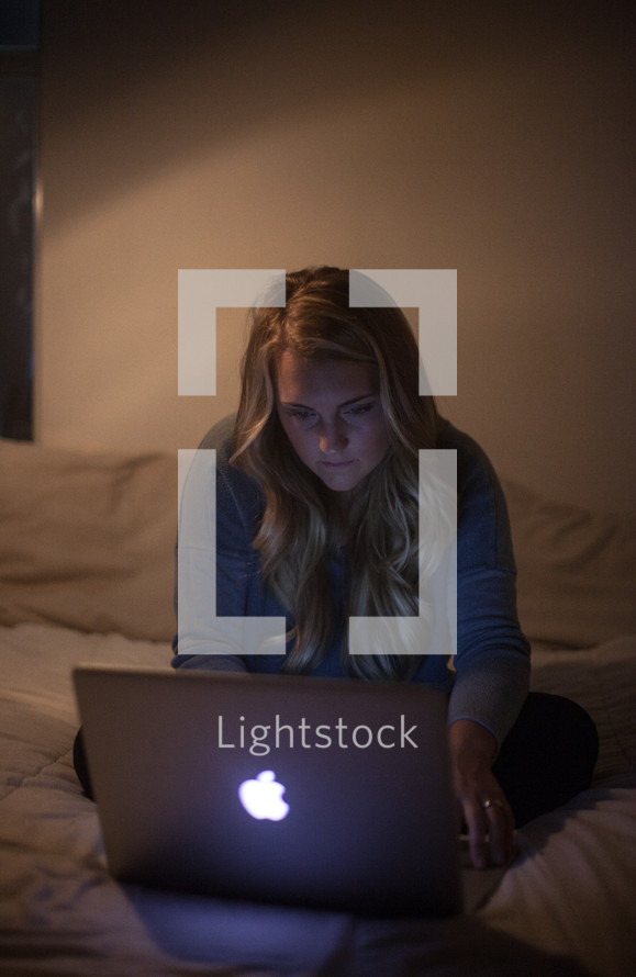 young woman sitting on her bed looking at a computer screen in a dark room 