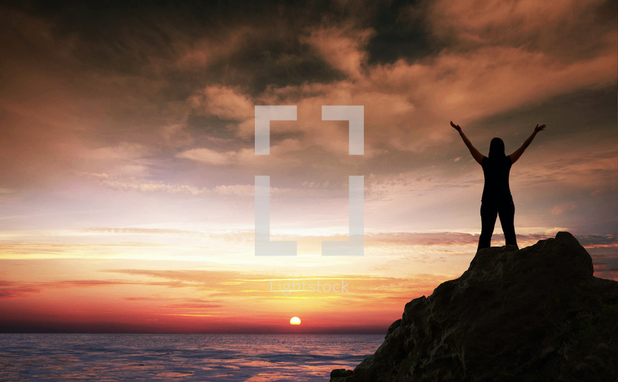 silhouette of a woman standing on a rock in front of the ocean at sunset with her hands raised in worship to God