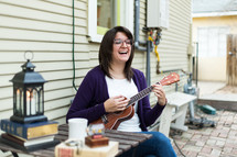 A smiling woman sits outside playing a ukulele.