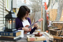 A young woman plays a ukulele at an outdoor table.