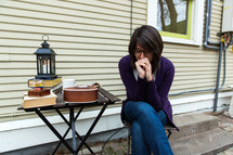A woman, with her head down in prayer, sits at an outdoor table full of books, lantern, and a ukulele.