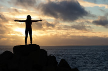 a woman standing on a rock by a shore with outstretched arms 
