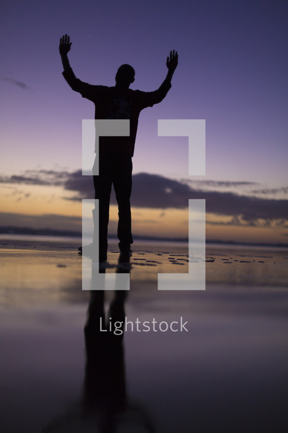 silhouette of a man standing on a beach with hands raised in worship 