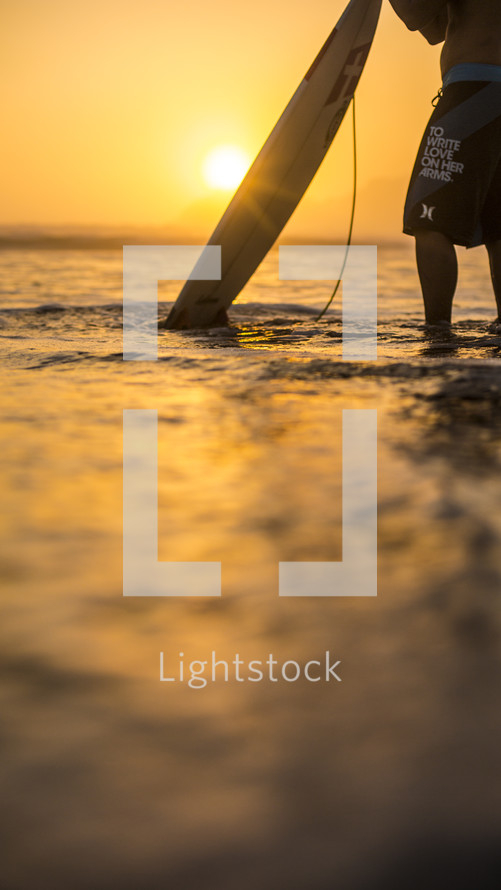 surfer holding his surfboard in the ocean at sunrise