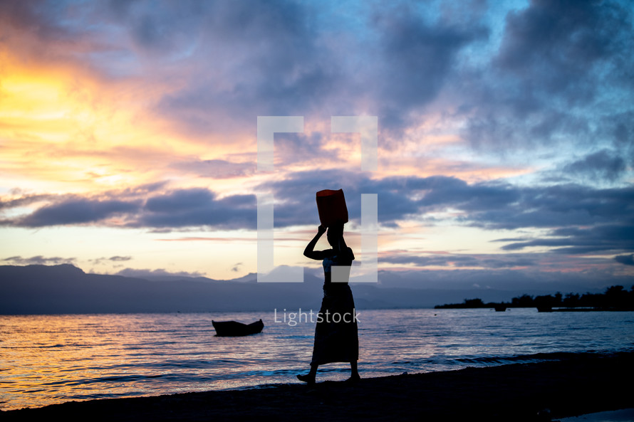 a woman walking on a beach at sunset with a basket on her head 