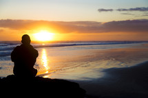 silhouette of a man sitting on a rock on a beach at sunset 