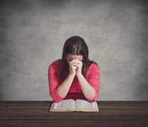 Woman praying over a Bible on a wooden table.