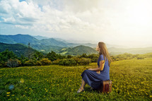Woman sitting on a suitcase in a grassy meadow with flowers, overlooking a mountain range.