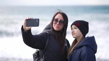 Cheerful young mother and daughter taking selfie on the beach. Smiling mother and daughter taking selfies at beach on a spring day. Family lifestyle concept.