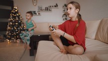 Video game concept. Pretty girl and little boy playing game console and laughing while sitting on sofa at home. Girl playing video games with a joystick. Child gambling addiction.