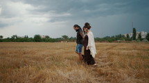Man in a black cloth and a pregnant woman in a black long dress and dreadlocks embrace and are kept in hand, against the background of yellow grass field, nature, autumn or summer. love story