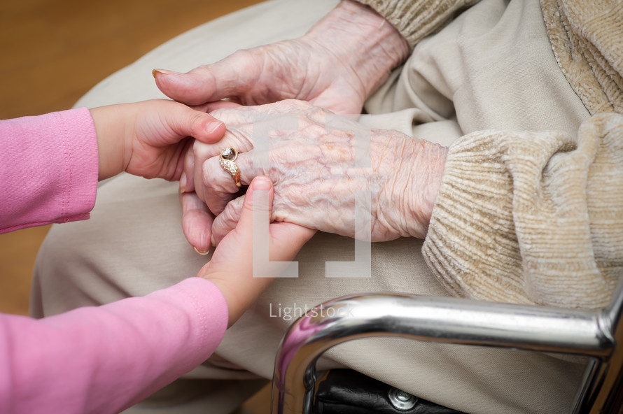 Child's hands holding the hands of a grandmother in a wheelchair.