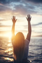 girl with raised hands standing in front of the ocean on a beach at sunset 