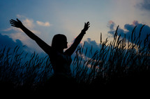 silhouette of a woman standing in a field with her arms raised to God