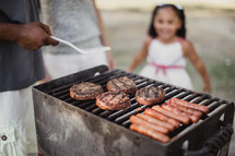 A little girl watches a man cooking hamburgers and hot dogs on an outside grill.