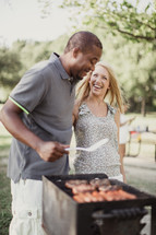 A man and woman laughing together while cooking on an outside grill.