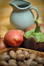 Still life with ceramic pitcher, basket of pecans, eggplant, and potatoes.

