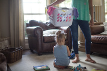 a mother holding a laundry basket with her daughter playing with toys on the floor 