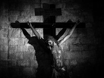 A black & white (b&w) crucifix, statue of Jesus, wrapped in a shawl against a stone brick wall.