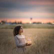 a girl standing in a field holding a jar of fireflies 