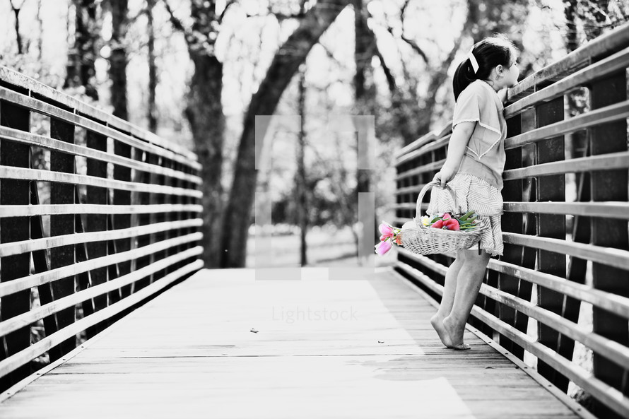 Girl carrying basket of flowers across wooden bridge.