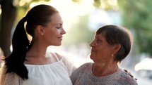 Beautiful portrait of grandmother and her granddaughter look at each other with love on summer street. Sun flares. Girl embracing granny with gratitude. Family concept. 4k