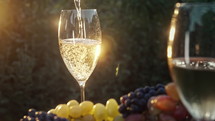 Stunning view of pouring white wine into glass goblet with fresh grape bunch background, sun light breaks through vineyard. 