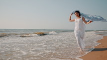 Woman in a white dress running along the shoreline of a beach.