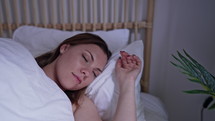 Young calm woman sleeping well alone in comfortable bed. Lady with happy face and smile in light cozy bedroom. Healthy resting in morning, enjoying dreaming