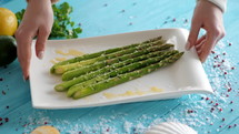 Female hands puts a plate of freshly prepared asparagus on a wooden blue table decorated with sea salt, herbs and spices. 