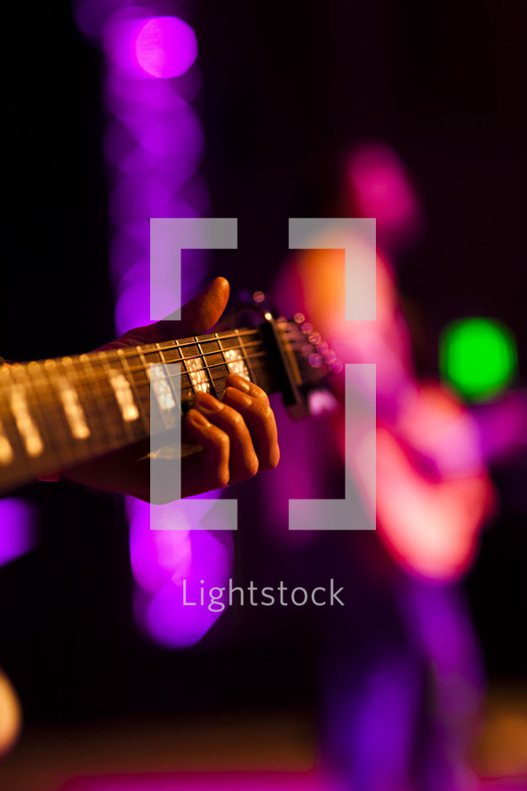 Hand holding guitar with other player in background on lighted stage fret board
