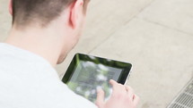 Man reading on an electronic table while standing outside on a sidewalk.