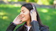 Happy mixed race young woman listening to music on headphones. Hispanic hipster girl dancing to rhythm and singing along melody in the park.