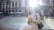  Pretty woman using smartphone on city street. Lady sitting alone on fountain in center square, she surfing internet with mobile device. Autumn season. Technology, social apps, connection concept. 4k

