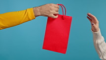 Woman gives gift bag to man. Female hand with red package on blue background. Concept of seasonal sale, purchases, shopping. High quality 4k footage