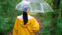 Young pretty woman in yellow raincoat walking in autumn park or forest alone. Girl with transparent umbrella. Rainy weather, nature background.